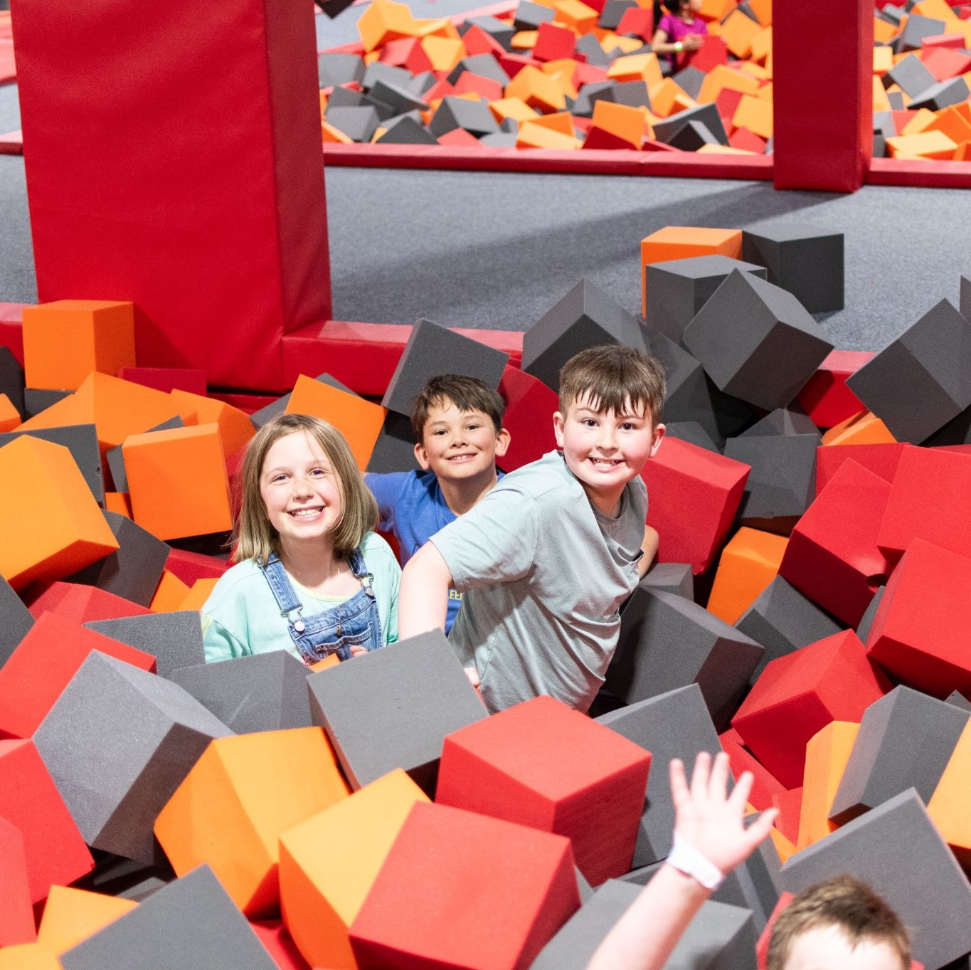 Children smiling and playing in a foam pit filled with red, orange, and gray cubes