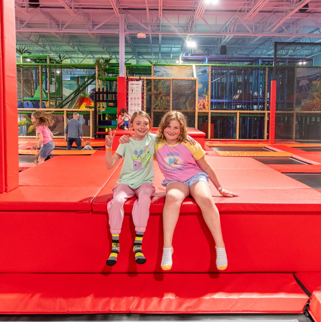 Two kids sitting and smiling on a red trampoline platform in an indoor play center